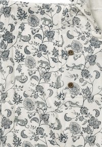 Blommönstrad tyg i vitt och mörkblått med lejon och blommor, med bruna knappdetaljer fram. Texturerat bomullsmaterial.