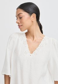 Blusa blanca con mangas cortas abullonadas, con un escote en forma de V adornado con bordados florales y un delicado acabado de encaje. Tejido texturizado.