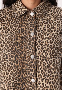 Spijkerjasje met luipaardprint, een kraag, zilveren drukknopen en twee borstzakken. Aardse tinten domineren het ontwerp.