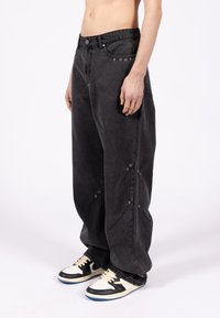 Czarne, luźne spodnie z denimu w stylu oversize, ozdobione zatrzaskami z przodu i po bokach, z szarym przeszyciem. W zestawie z czarno-białymi trampkami.