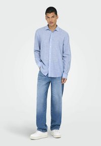 Camicia azzurro chiaro a maniche lunghe con colletto a bottoni, abbinata a jeans blu a vestibilità morbida e sneakers bianche, in piedi contro uno sfondo neutro.