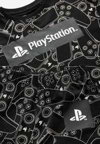 Tissu noir avec un motif blanc de manettes de jeu. Présente une étiquette centrale grise avec le logo « PlayStation » et une étiquette. Texture douce.
