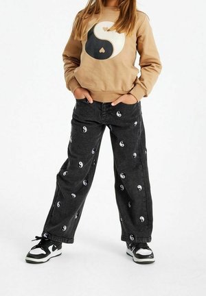 Dziecko ubrane w beżową bluzę z dużym symbolem yin-yang oraz czarne spodnie w mniejsze symbole yin-yang, do tego czarno-białe sneakersy.