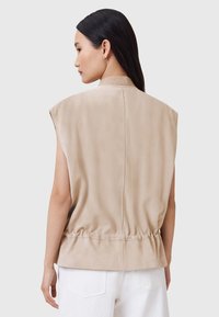 Gilet senza maniche beige realizzato in morbido suede, con vita sfiancata, colletto alto e una texture liscia. Dettagli di cucitura discreti sul retro.