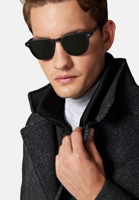 Czarne okulary przeciwsłoneczne z ciemnymi soczewkami, noszone przez osobę w fakturowanym czarnym płaszczu i białym golfie. Kołnierz jest częściowo zapięty.