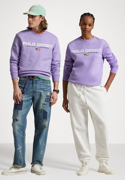 Dva modely majú na sebe fialové mikiny s nápisom "POLO SPORT". Jeden má na sebe poškodené džínsy s nášivkami, druhý biele tepláky a tenisky.