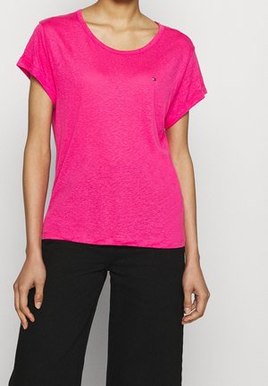 Różowy t-shirt z krótkim rękawem i okrągłym dekoltem oraz małym logo na piersi. Miękki, teksturowany materiał, luźny krój, zestawiony z czarnymi spodniami.