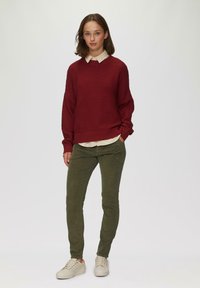 Bordeauxfarbenes, geripptes Sweatshirt mit überschnittenen Schultern, über einem hellbeigen Hemd mit Kragen getragen. Olivgrüne, schmal zulaufende Hose und cremefarbene Sneaker.