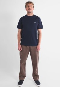 T-shirt en coton bleu marine à manches courtes et col rond, avec un petit logo sur la poitrine, associé à un pantalon marron clair à coupe décontractée.