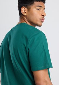 Green cotton t-shirt with short sleeves, crew neck, and a smooth texture. The model is seen from a profile angle, showcasing the shirt's fit.