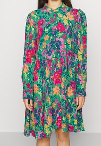 Blumenkleid mit grünem Grundton, gepunktet mit rosa, orangefarbenen und gelben Blumen, langen Ärmeln, rundem Kragen und einem lockeren, ausgestellt geschnittenen Design.
