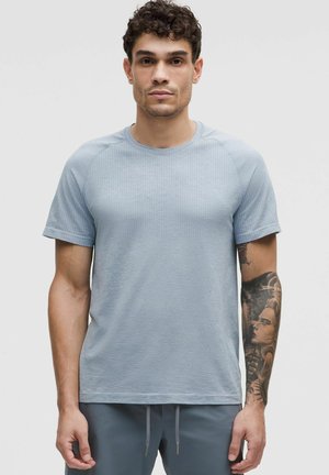 T-shirt azzurro chiaro a maniche corte con motivo testurizzato, scollatura rotonda e maniche raglan, indossato con pantaloni grigi e con tatuaggi visibili sul braccio.