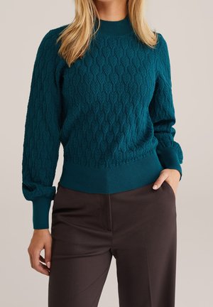 Femme portant un pull en maille torsadée bleu sarcelle foncé à col montant et un pantalon droit marron foncé, une main dans la poche, fond clair.