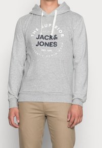 Sudadera gris con capucha ajustable, que cuenta con un bolsillo tipo canguro en la parte delantera y un logotipo gráfico circular en azul oscuro. Textura de tela suave.