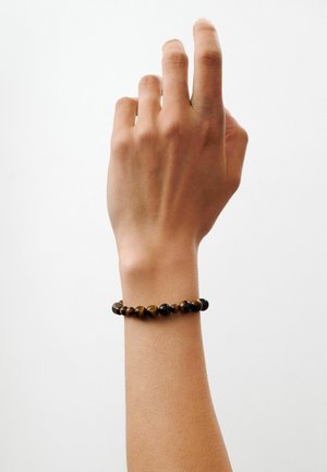 Hand met een medium huidtint die een bruin-zwart kralenarmband draagt, opgeheven tegen een effen witte achtergrond.