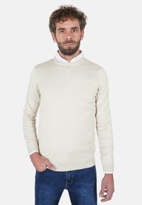 Uomo adulto con capelli ricci e barba che indossa un maglione beige sopra una camicia bianca e jeans blu, in piedi con una mano in tasca.