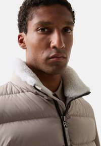 Beige dunjakke med hvid imiteret pelskrave, horisontal syning og en lynlåsedetalje med en metaltrækflig.