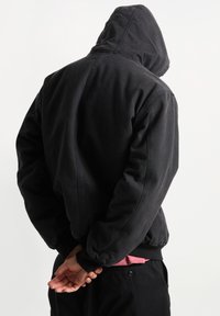 Giacca nera con cappuccio, dal taglio rilassato, realizzata in un tessuto texturizzato, con polsini a costine e cappuccio con cordino. Design minimalista, senza loghi visibili.