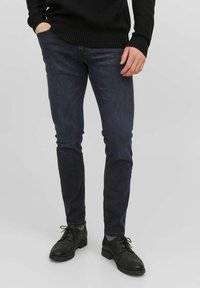 Man med mörkblå slim-fit jeans, svart stickad tröja, grå strumpor och svarta läder snörskor, som står med en hand i fickan.