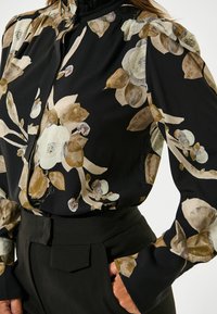 Schwarz-bluse mit Blumenmuster in Beige und Braun, hohem Kragen und lockeren Ärmeln. Der Stoff wirkt leicht und fließend.