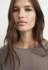 Grå, texturerad stickad topp med vertikala ribbor, bred halsringning och mjuk fall. Modellen har lång brun hår och naturlig sminkning.