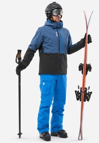 Skiausrüstung mit einer blauen und schwarzen wasserdichten Jacke, blauen gemusterten Hosen, schwarzen Handschuhen, einem Skistöck und Skiern mit roten Akzenten.