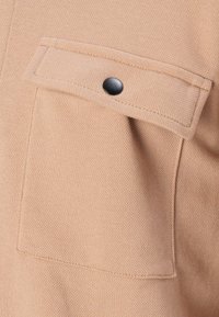 Gros plan sur une poche en tissu beige avec un rabat sécurisé par un seul bouton-pression noir sur un matériau texturé.