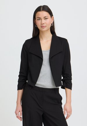 Kobieta ubrana w czarny krótki żakiet na jasnoszarym topie i czarne spodnie, stojąca na tle jednolitego białego tła.