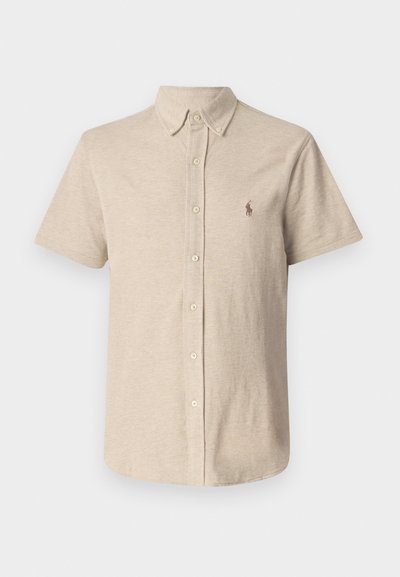 Camisa de botones de manga corta en color beige, con un cuello clásico y un pequeño logo bordado en el lado izquierdo del pecho. Fabricada en tela de algodón texturizada.