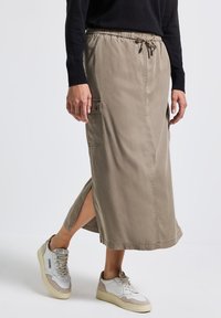 Henkilö, jolla on päällään beige midi-hame, jossa on sivutaskut ja halkio, musta pitkähihaine paita sekä valkoiset lenkkitossut, joissa on beigeä korosteväriä.