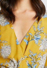 Gelbes Wickelkleid mit blauen und weißen Blumenmustern, V-Ausschnitt und weicher, glatter Textur.