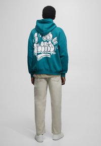 Persona che indossa una felpa teal con graffiti bianchi sulla schiena, pantaloni beige e sneakers bianche, in piedi davanti a uno sfondo grigio chiaro uniforme.