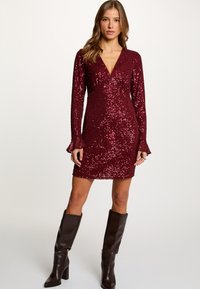 Viininpunainen paljettimekko, jossa pitkät hihat, syvä V-pääntie ja levenevät kalvosimet. Yhdistetty polven korkeisiin ruskeisiin korkosaapaisiin. Sileä pinta ja istuva malli.