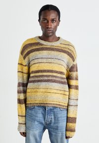 Pull rayé aux couleurs jaune, marron et gris avec une finition douce et texturée. Encolure ronde côtelée et manches longues. Design court.