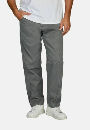 Mann trägt verblasste graue Hose mit geradem Bein und weiße Sneaker, steht mit einer Hand in der Tasche vor einem schlichten Hintergrund.