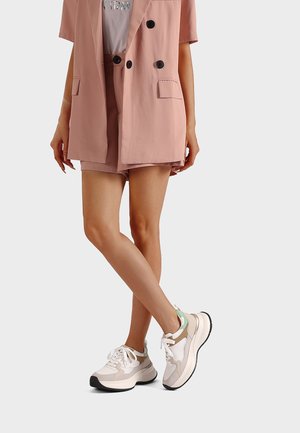 Donna con blazer rosa e pantaloncini abbinati a sneaker chunky bianche e beige, in piedi con le gambe incrociate su uno sfondo chiaro.