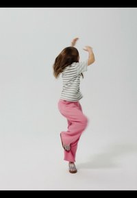 Child with long brown hair wearing a striped shirt and pink pants jumping with arms raised against a plain white background.