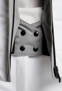 Grå och vit vadderad jacka med svarta tryckknappar, kontrasterande rosa foder och en synlig dragkedja. Mjuk interiörtextur.