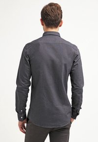Chemise à manches longues gris foncé avec col à boutons, texture lisse, empiècement dos, et poignets à un bouton. Coupe ajustée avec ourlet arrondi.