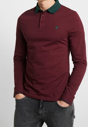 Homme portant un polo manches longues bordeaux avec un col vert foncé et un logo brodé, mains dans les poches, assorti à un jean gris foncé.