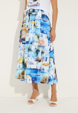 Frauen, die einen Midirock mit einem mediterranen Küstendorf-Print tragen, kombiniert mit weißen offenen High Heels, von der Taille bis zu den Füßen gezeigt.