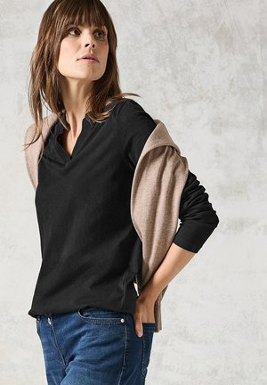 Svart långärmad topp med V-ringning, lager under en ljusbrun stickad sjal, kombinerad med blå jeans, på en texturerad ljus bakgrund.