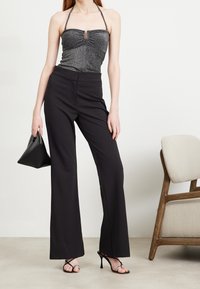 Kobieta ubrana w czarne spodnie o szerokich nogawkach, błyszczący top na halter, czarne sandały na wysokim obcasie, trzymająca czarną torebkę, stojąca obok beżowego krzesła.