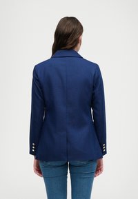 Navy blazer gemaakt van gestructureerde stof, met een gelapt kraag, een getailleerd ontwerp en gouden knopenaccenten op de mouwen.