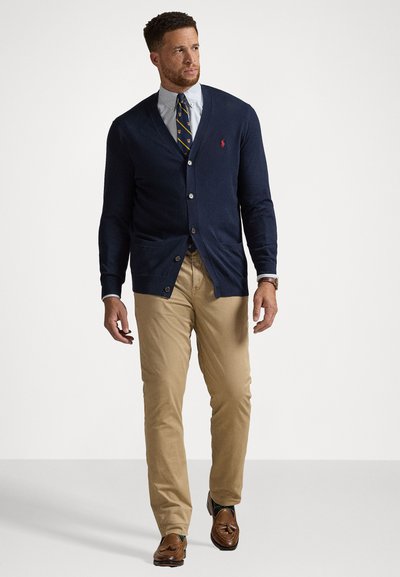 Cardigan din amestec de lână navy cu închidere cu butoane, două buzunare frontale, combinat cu o cămașă bleu deschis și o cravată cu model, pantaloni chinos bej, mocasini maro.