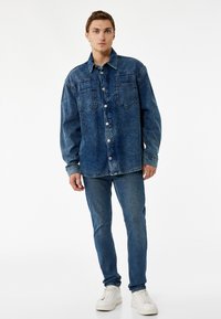 Camisa de mezclilla en azul oscuro con mangas largas, dos bolsillos en el pecho y cierre de botones. Lleva puesta sobre unos jeans ajustados de mezclilla y zapatillas blancas.
