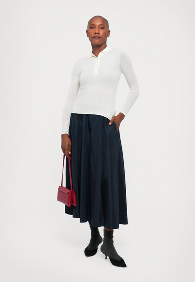 Polo bianco a maniche lunghe con colletto, abbinato a una gonna plissettata blu navy. Accessoriato con una borsa crossbody rossa e décolleté nere a punta.
