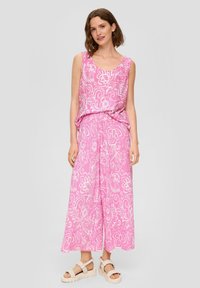 Roze, mouwloze jumpsuit met witte bloemmotieven, losse pasvorm en een getailleerde taille. Gecombineerd met gevlochten plateau-sandalen.