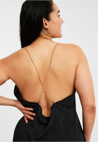 Schwarzes Kleid mit tiefem Rückenausschnitt und über Kreuz gelegten goldenen Ketten als Träger, das eine glatte, fließende Textur und weichen Stoff aufweist.