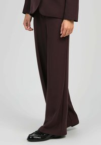 Personne portant un pantalon palazzo marron foncé, un blazer assorti et des mocassins noirs brillants, se tenant devant un fond clair uniforme.